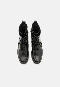 Botas de tobillo de cuero negro con acabado brillante, diseño con cordones, cremalleras laterales y una correa decorativa con detalle de hebilla de metal.