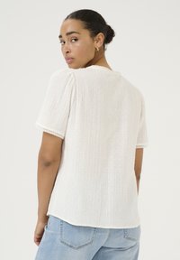 Blouse blanche texturée à manches courtes et dentelle, avec des motifs en relief et un col rond, assortie à un jean bleu clair.