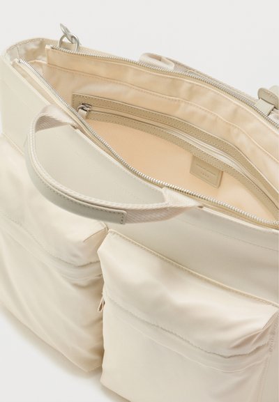Τσάντα χειρός σε κρεμ χρώμα με δύο μπροστινές τσέπες με φερμουάρ, υφασμάτινα χερούλια και έναν κύριο χώρο με εσωτερική τσέπη με φερμουάρ.