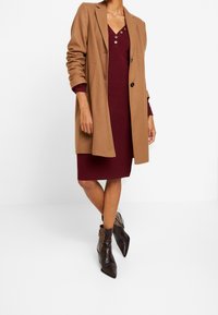 Un manteau long couleur camel porté sur une robe bordeaux mi-longue avec un col à boutons. Des bottines à talons en cuir marron à bout pointu avec une texture en cuir serpent.