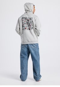 Gray hoodie with a graphic design on the back, featuring colorful patterns. The model wears loose blue denim jeans.