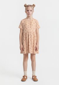 Fiatal lány semleges arckifejezéssel, barackvirágos rövid ujjú ruhát, krémszínű zoknit és barna cipőt visel, haját két kontyba fonták.