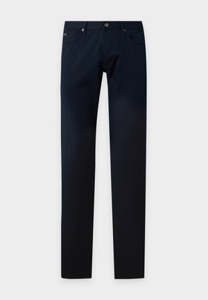 Uomo in piedi che indossa una polo beige chiaro a maniche corte, pantaloni blu scuro e sneaker bianche su uno sfondo bianco semplice.