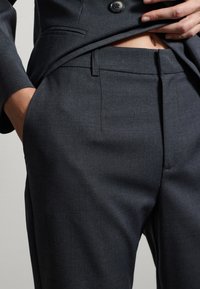 Gros plan sur une personne portant un costume sur mesure gris foncé, avec une main dans la poche et l'autre soulevant la veste pour révéler la taille.