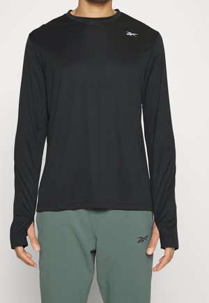 Man draagt een zwart sportshirt met lange mouwen en duimgaten en een groene broek, beide met Reebok-logo's.
