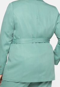 Blazer verde menta con un design su misura, texture liscia, e un nastro in tessuto in vita, con spalle e maniche strutturate.