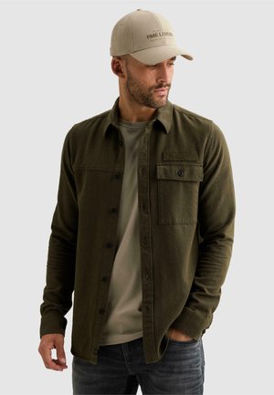Homme portant une casquette beige, une chemise verte olive à boutons par-dessus un T-shirt gris clair, et un jean foncé, regardant vers le bas et sur le côté avec une main dans la poche.