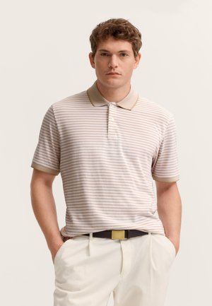 Junger Mann mit kurzen lockigen Haaren, der ein beige-weiß gestreiftes Poloshirt und weiße Hosen trägt, steht mit den Händen in den Taschen.