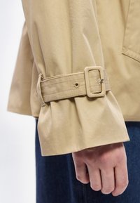 Main portant une veste beige avec une sangle à boucle réglable au niveau du poignet et un pantalon bleu foncé visible en arrière-plan.