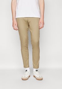 Pantalons slim-fit beiges en tissu lisse, avec une coupe fuseau. Associés à des baskets blanches et des chaussettes blanches visibles.