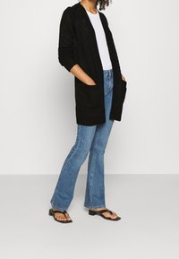 Žena oblečená v černém pleteném kardiganu, bílém topu, modrých rozšířených džínách a černých sandálech na vysokém podpatku, stojí s rukama v kapsách.