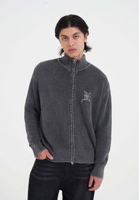 Junger Mann mit schulterlangen, dunklen Haaren, der einen grauen Reissverschluss-Pullover mit Rippenstruktur und schwarze Hosen trägt, steht vor einem schlichten weißen Hintergrund.