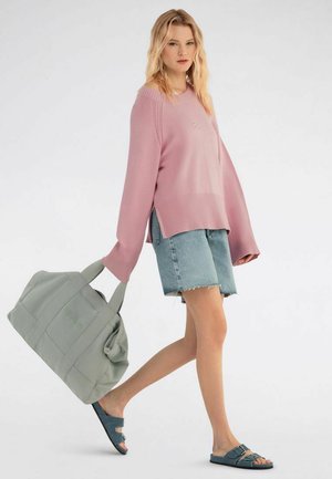 Junge Frau geht spazieren, trägt einen übergroßen rosa Pullover, Jeansshorts, blaue Sandalen und hält eine große graue Reisetasche in ihrer linken Hand.
