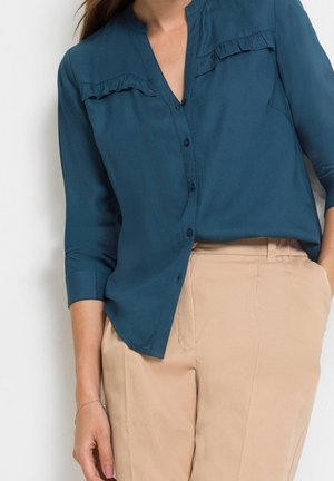 Kobieta ubrana w turkusową bluzkę zapinaną na guziki z falbanką i beżowe spodnie z wysokim stanem, stojąca przy białej ścianie.