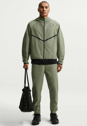 Homme portant un survêtement Nike vert olive et des baskets noires, tenant un sac fourre-tout noir avec des poignées en corde, debout sur un fond uni.