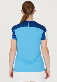 Person med hästsvans som bär en kortärmad blå sporttröja med vita och mörkblå randiga ärmar, vänd bort från kameran mot en enkel bakgrund.