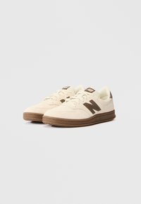 Sneakers w kolorze kremowym, wykonane z zamszu, z brązowymi akcentami i teksturowaną gumową podeszwą. Klasyczny design z perforowanymi detalami na palcach.
