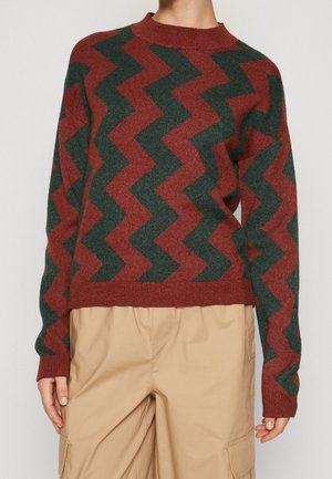 Person iført en rustrød og mørkegrøn zigzag-mønstret sweater med beige cargobukser, stående mod en ensfarvet lys baggrund.
