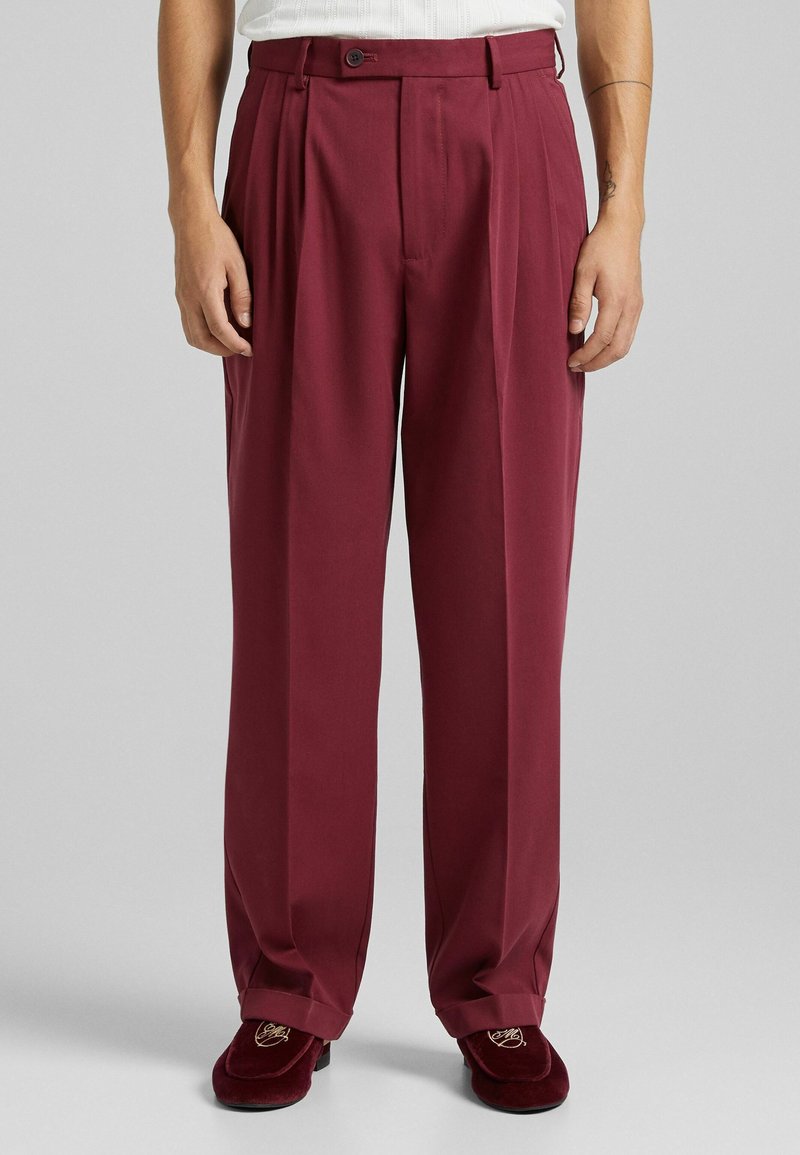 Pantaloni plissettati bordeaux con vita alta, realizzati in un tessuto liscio. Presentano una chiusura con un bottone e tasche laterali. Vestibilità sartoriale.
