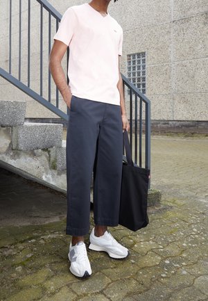 T-shirt rose clair en V, pantalon large bleu marine et baskets blanches avec accents gris. Sac tote noir tenu à la main, escaliers en béton en arrière-plan.
