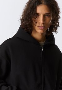 Felpa nera con zip, realizzata in tessuto morbido, caratterizzata da un grande cappuccio, vestibilità comoda e dettaglio a zip a contrasto sul davanti.