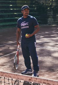 Jogador de ténis com uma camisola azul-marinho com um gráfico rosa e o texto "LACOSTE", calças azul-marinho com detalhes brancos, segurando uma bola de ténis e uma raquete.
