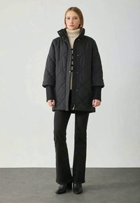 Sort quilted dunjakke med høj krave, oversized ærmer og lynlås foran, parret med en cremefarvet turtleneck og sorte flagrende bukser.