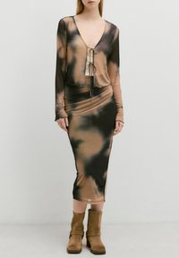 Langärmliges Kleid mit Schleifenverschluss in einer Mischung aus braunen und schwarzen Batikmustern, aus einem dehnbaren, glatten Stoff gefertigt, kombiniert mit braunen Stiefeletten.