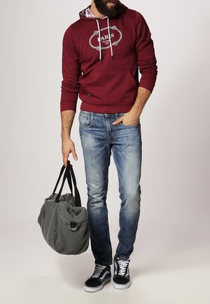 Man draagt een kastanjebruwe hoodie met de tekst "Paris", blauwe spijkerbroek, zwarte sneakers en houdt een grijze weekendtas bij de handvatten vast.