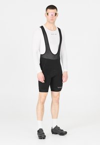 Cuissard de cyclisme pour homme, noir avec des bretelles en mesh, doté d'un rembourrage à l'assise. Sous-vêtement à manches longues blanc, lunettes de soleil miroir roses, chaussures de cyclisme noires.