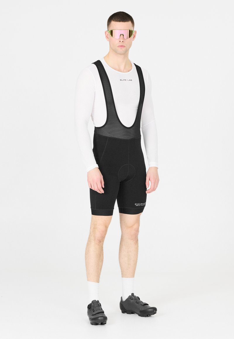 Cuissard de cyclisme pour homme, noir avec des bretelles en mesh, doté d'un rembourrage à l'assise. Sous-vêtement à manches longues blanc, lunettes de soleil miroir roses, chaussures de cyclisme noires.