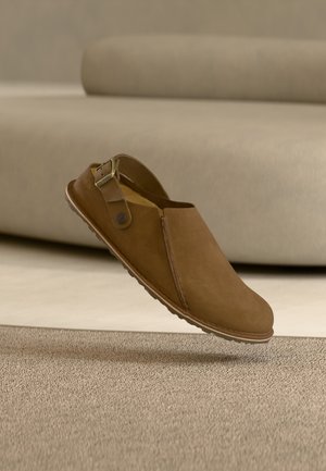 Brauner Wildleder-Slipper-Clog mit verstellbarem Lederband und Gummisohle, schwebt über einem beigen Teppich in einer neutralen Innenraumumgebung.