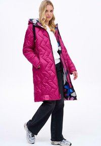 Růžový vypolstrovaný dlouhý kabát s kapucí, vyznačující se barevnou květinovou podšívkou. Doplněný o černé kalhoty a sportovní boty.