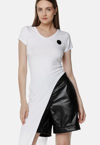 Biała koszulka z dekoltem w serek i krótkimi rękawami, ozdobiona czarnym okrągłym logo, w połączeniu z czarnymi, błyszczącymi szortami do kolan z podwiniętymi mankietami.