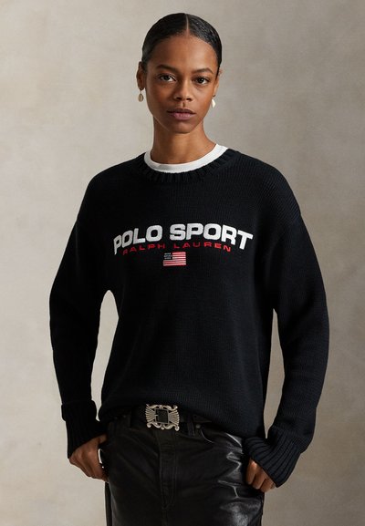 Sileäksi kammatut hiukset omaava nainen pukeutunut mustaan Polo Sport Ralph Lauren -neuleeseen ja mustiin nahkahousuihin, joissa koristeellinen vyönsolki, yksiväristä taustaa vasten.