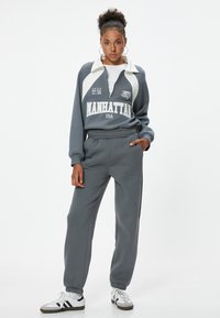 Grijze tweedelige trainingsoutfit bestaande uit een rits-vest met een witte kraag en logo, gecombineerd met bijpassende taps toegesneden trainingsbroek.