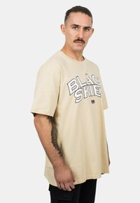 Beige pamut póló, melyen vastag fekete betűkkel szerepel a "BLACK SKIES" felirat. Laza szabású, rövid ujjú és minimalista design.