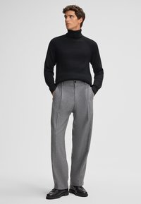 Maglione a collo alto nero a costine abbinato a pantaloni grigi chiari con pieghe. Il modello indossa scarpe nere. Design semplice e contemporaneo, senza motivi visibili.