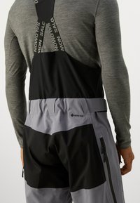 Salopete izolate GORE-TEX în negru și gri, dotate cu bretele ajustabile, fermoare laterale și un material texturat pentru flexibilitate și durabilitate.
