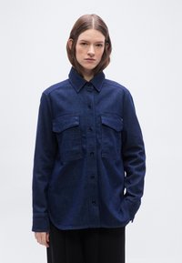 Νεα ρή γυναίκα με ίσια καστανά μαλλιά που φορά σκούρο μπλε τζιν πουκάμισο με κουμπιά και δύο τσέπες στο στήθος, στέκεται μπροστά σε απλό φόντο.