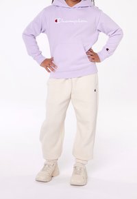Sudadera de lavanda con logo "Champion" blanco, bolsillo frontal y puños acanalados; combinada con pantalones de chándal crema y zapatillas beige.