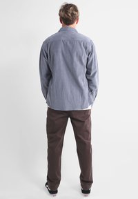 Camisa gris de botones con una tela texturizada, combinada con pantalones marrones y zapatillas negras. La camisa cuenta con un cuello clásico y pliegues en la espalda.