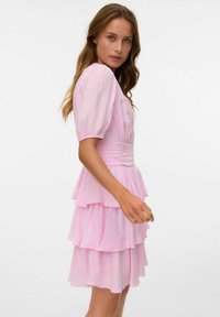 Rosa tierad klänning med puffärmar, texturerad tyg och figursydd midja. Har tre volangskikt och en mjuk, flödande silhuett.