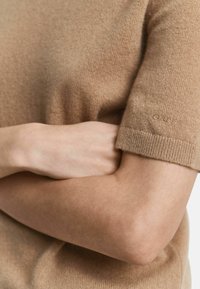 Die Arme vor der Brust verschränkt, mit einem kurzärmligen, beigen Strickpullover, auf dem das GANT-Logo auf dem Ärmel gestickt ist.