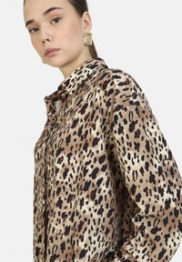 Camicia in satin con stampa leopardata e colletto, caratterizzata da una texture morbida in tonalità di beige, marrone e nero. Dettagli con chiusura a bottoni visibile.