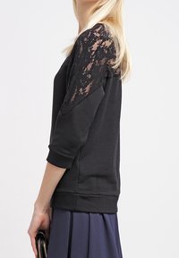 Sweat-shirt noir avec manches en dentelle trois-quarts et encolure ronde. Texture douce avec une coupe ajustée, associé à une jupe plissée.