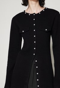 Cárdigan negro de punto con botones de perlas en el frente, cuello y bolsillos con solapa, llevado sobre pantalones negros.