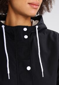 Chaqueta negra con capucha, cordones blancos y botones a presión. Fabricada con un tejido suave y con un forro interior suave.