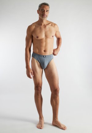 Man met kort grijs haar, gekleed in grijze slip, staand op blote voeten tegen een witte achtergrond, met één hand op zijn heup.