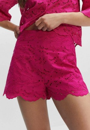 Femme ajustant un haut en dentelle fuchsia assorti à un short en dentelle fuchsia avec des bords festonnés et des motifs découpés floraux.